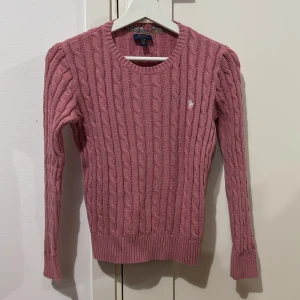 Rosa stickad tröja från Ralph Lauren kids - Säljer en snygg rosa stickad tröja från Ralph Lauren med kabelstickat mönster. Tröjan har långa ärmar och en rund halsringning. Tröjan är i väldigt bra skick.