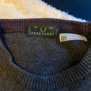 Grå tröja från Fred Perry - Säljer en stilren grå tröja från Fred Perry med rund halsringning och ribbade muddar. Tröjan har en diskret lila logga broderad på bröstet. Perfekt för en avslappnad och snygg look.