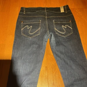 Mörkblå jeans med broderade bakfickor - Raka, lågmidjade, mörkblå jeans med broderade detaljer på bakfickorna. De har en klassisk femficksdesign och knappar framtill. 