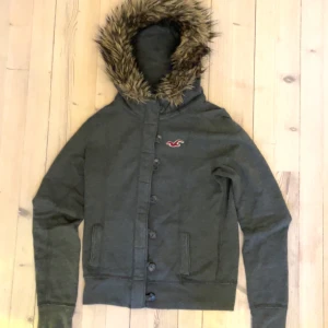 hoodie med pälskrage från Hollister - Säljer en snygg grå jacka från Hollister med en mysig pälskrage och knappar framtill. Tror att de är tjej storlek 