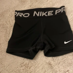 Svarta träningsshorts från Nike - Säljer ett par svarta Nike Pro träningsshorts med en elastisk midja och vit logga. Perfekta för intensiva träningspass eller löpning. De är designade för att sitta tight och ge stöd under aktivitet.