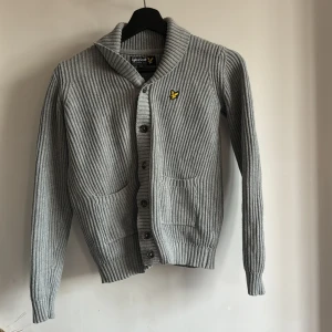 Grå ribbad kofta från Lyle & Scott - Säljer en stilren grå ribbad kofta från Lyle & Scott med knappar framtill och två fickor. Den har en broderad logotyp på bröstet och långa ärmar med ribbade muddar. Perfekt för kyligare dagar! Passar stl 152