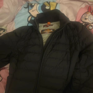 Svart dunjacka från Parajumpers - Säljer en svart dunjacka från Parajumpers med dragkedja och hög krage. Jackan har ett quiltat mönster och en broderad logga på ärmen. Perfekt för kyliga dagar!