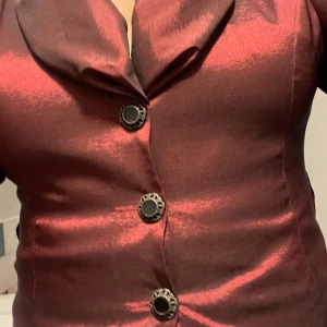 Vinröd kavaj med dekorativa knappar - Elegant vinröd kavaj med glänsande yta och dekorativa svarta knappar. Kavajen har en figurnära passform och trekvart  ärmar. Perfekt för en stilfull look.