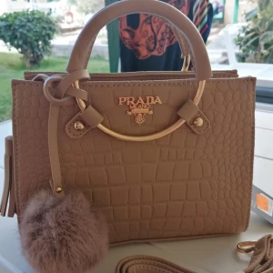 Beige handväska från Prada - Snygg beige handväska från Prada med krokodilpräglat mönster och gulddetaljer. Väskan har en rund handtag och en fluffig pompom som dekoration. Perfekt för att ge en lyxig touch till din outfit, den är även avtagbar.