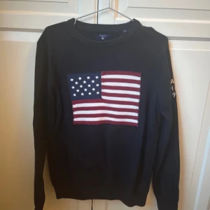 Tröja med amerikansk flagga från GANT - Säljer en tröja från GANT med en stor amerikansk flagga på framsidan. Tröjan har långa ärmar och texten 'GANT NH-CT 1949' på ärmen. den är mörkblå/marinblå