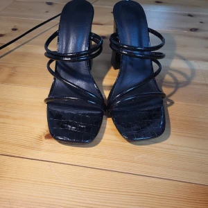 Svarta klackskor med remmar - Snygga svarta klackskor med flera tunna remmar och en fyrkantig tå. Skorna har en hög klack och en krokodilpräglad yta som ger en lyxig känsla. Perfekta för en kväll ute eller en speciell tillställning. P.S skorna har bara blivit använda en gång.