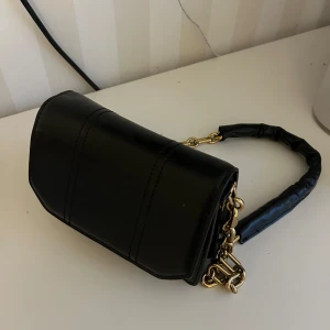 Svart axelväska med kedja - Snygg svart axelväska i skinn med guldfärgad kedja som axelrem. Väskan har en praktisk dragkedja och en knappstängning framtill. Aldrig använd, så tänker kan komma till nytta hos någon annan!☺️