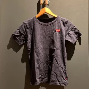 Svart t-shirt från Levi's - Säljer en klassisk svart t-shirt från Levi's med deras ikoniska röda logga på bröstet. T-shirten är tillverkad i 100% bomull och har en rund halsringning. Perfekt för en avslappnad stil.