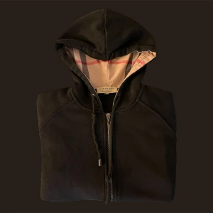 Svart hoodie från Burberry - Säljer en stilren svart hoodie från Burberry med dragkedja och klassiskt rutigt foder i huvan. Perfekt för en avslappnad och trendig look. Huvan har justerbara snören och tröjan har en praktisk ficka framtill.
