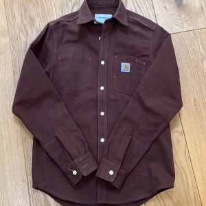 Brun skjorta från Carhartt - Snygg brun skjorta från Carhartt med klassisk krage och vita knappar. Skjortan har två bröstfickor med Carhartt-logga på ena fickan. Perfekt för en stilren look.