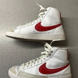 Vita och röda Nike sneakers - Snygga vita Nike sneakers med röd swoosh och hög modell. Skorna har snörning och en klassisk design som passar perfekt för en sportig look. Tillverkade i syntetmaterial med en bekväm passform. Sparsamt använda! 