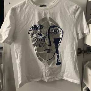 Zara tröja  - Säljer en vit t-shirt från Zara med ett unikt konstnärligt ansiktstryck i blått och sliver på framsidan. Perfekt för dig som vill ha något stilrent. 