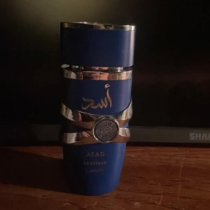 Asad Zanzibar parfym från Lattafa - Elegant parfymflaska i djupblått med silverdetaljer och arabiska inskriptioner. Flaskan har en unik form och en dekorativ silverring runt mitten. Perfekt för den som söker en distinkt och lyxig doftupplevelse.