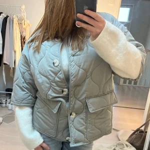  jacka med korta ärmar - Säljer en stilren grå quiltad jacka med stora knappar och fickor framtill. Jackan har en oversized passform och trekvartsärmar, perfekt för lager-på-lager-stil. Den quiltade designen ger en trendig touch. Perfekt för kyligare dagar!
