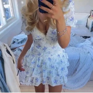 Blommig vit klänning med volanger - Charmig vit klänning med blå blommönster och volangdetaljer. Klänningen har en v-ringad design och korta ärmar, perfekt för sommarens festligheter.