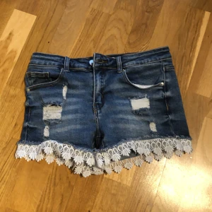 Blå jeansshorts med spets från Hailys - Snygga blå jeansshorts från Hailys med slitna detaljer och vit spetskant längst ner. Perfekta för en somrig look. De har en klassisk femficksdesign och knappgylf.