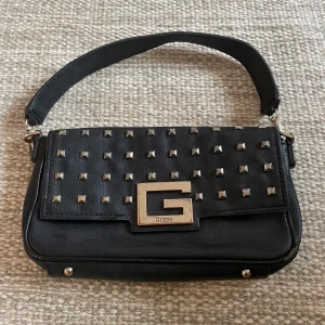 Svart axelväska från Guess - Svart väska från Guess med nitar, lång och kort band medkommer. Köpt för 1000kr säljer för 400
