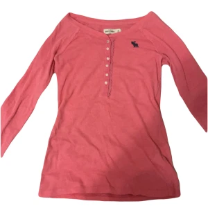 Rosa långärmad topp från Abercrombie - Säljer en söt rosa långärmad topp från Abercrombie med knappar framtill och ett litet mörkblått logotryck på bröstet. Perfekt för en avslappnad stil. 💖 M i barnstorlek men passar xs och formar kroppen bra. Defekter är att tröjan är nopprig annars finns det inget mer fråga för bilder på