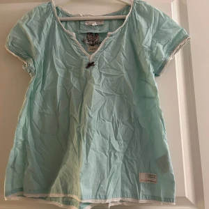 Ljusblå kortärmad blus från Odd Molly - Säljer en söt ljusblå blus från Odd Molly med korta ärmar och v-ringning. Toppen har en liten rosett framtill och dekorativa spetsdetaljer vid halsen, kanten och ärmsluten. Perfekt för en avslappnad stil. Blusen är i fint och välvårdat skick. Vid frågor och funderingar hör av er, jag svarar så fort jag kan!🩷