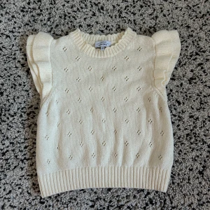 Vit stickad topp från & Other Stories - Säljer en söt vit stickad topp från & Other Stories med volangdetaljer på axlarna. Toppen har ett diskret hålmönster och ribbade kanter vid hals och nederkant. Perfekt för en stilren look! 🌟