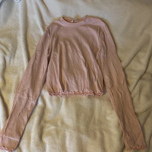 Rosa ribbad tröja från H&M x Hello Kitty - Säljer en söt rosa ribbad tröja från H&M i samarbete med Hello Kitty. Tröjan har långa ärmar och en glittrig Hello Kitty-design framtill. Den har en croppad passform med volangdetaljer vid nederkanten.