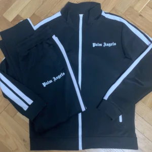 Palm angels & tracksuits - Snyggt svart träningsset från Palm Angels med vita ränder längs ärmar och ben. Jackan har dragkedja och hög krage, medan byxorna har en bekväm passform. Perfekt för en sportig look eller avslappnad stil.
