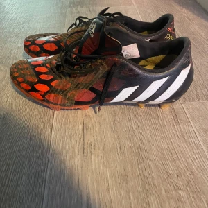 Adidas predator  - Säljer ett par Adidas fotbollsskor med ett unikt mönster i rött och svart. Skorna har snörning och är designade för att ge bra grepp på planen. Perfekta för fotbollsspelare som vill sticka ut.