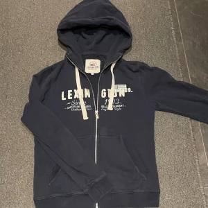 Mörkblå hoodie från Lexington - Säljer en super nice mörkblå hoodie från Lexington med dragkedja och tryck framtill. Den har en justerbar huva med vita snören