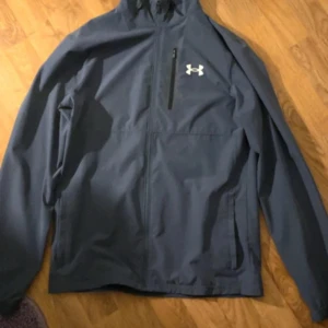 Blå vindjacka från Under Armour - Säljer en stilren blå vindjacka från Under Armour med dragkedja och huva. Jackan har en praktisk bröstficka med dragkedja och två sidofickor. Perfekt för blåsiga dagar och sportiga aktiviteter. Kan gå ner i pris vid snabb affär 