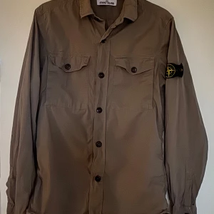Beige skjorta från Stone Island - Snygg beige skjorta från Stone Island med långa ärmar och klassisk krage. Skjortan har knappar framtill och två bröstfickor med lock. Den ikoniska Stone Island-loggan sitter på ärmen. Perfekt för en stilren look.