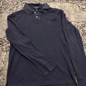 Mörkblå långärmad pikétröja från Ralph Lauren - Säljer en stilren mörkblå långärmad pikétröja från Ralph Lauren med klassisk krage och knappar framtill. Tröjan har det ikoniska röda broderade logotypen på bröstet. Perfekt för en avslappnad men ändå elegant look.