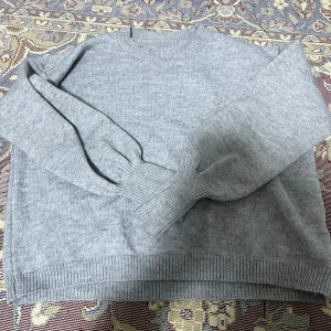 Grå stickad tröja - Säljer en s grå stickad tröja med rund hals och långa ärmar. Tröjan har ribbade muddar vid ärmslut och nederkant, vilket ger en klassisk look. Perfekt för kyligare dagar. Ganska skön att ha på sig !