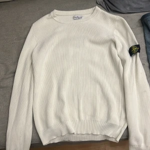 Vit tröja från Stone Island Junior - Säljer en stilren vit tröja från Stone Island Junior. Tröjan är ribbad och har långa ärmar med en ikonisk logga på ärmen. Perfekt för en avslappnad och trendig look.