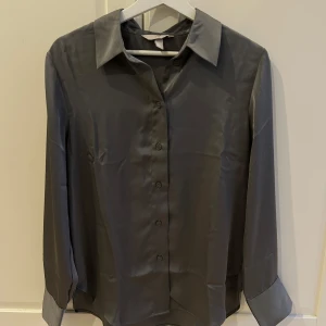 Grå skjorta från H&M - Elegant grå skjorta från H&M i ett glansigt material. Skjortan har långa ärmar och knappar framtill, vilket ger en klassisk look. Perfekt för en stilren och sofistikerad stil.
