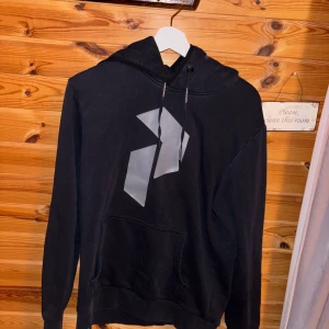 Svart hoodie från Peak Performance - Säljer en svart hoodie från Peak Performance med en stor ficka framtill och en stilren logga i grått på bröstet.  Nån fläck på ryggen men inget som inte går bort i tvätten😁