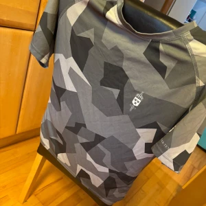 Grå kamouflage t-shirt från Försvarsmakten - Snygg grå kamouflage t-shirt från Försvarsmakten med korta ärmar. Perfekt för en cool och avslappnad stil. T-shirten har en modern design med texten 'FÖRSVARSMAKTEN' på ryggen. Storlek M