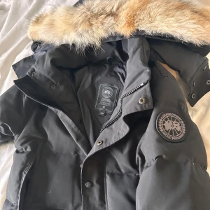 Svart parkajacka med pälsdetalj - Säljer en svart parkajacka från Canada Goose med en snygg pälsdetalj på huvan. Jackan har dragkedja och knappar framtill samt långa ärmar. Perfekt för kalla vinterdagar.