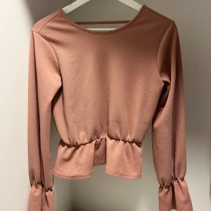 Rosa topp från NLY Trend - Snygg rosa topp från NLY Trend med volangdetaljer och v-ringad rygg. Toppen har långa ärmar med knytning vid ärmsluten och en elastisk midja för en smickrande passform.