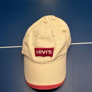 Vit keps från Levi's - Snygg vit keps från Levi's med röd broderad logga framtill. Kepsen har en justerbar rem baktill för perfekt passform och en röd kant på skärmen som ger en cool kontrast.