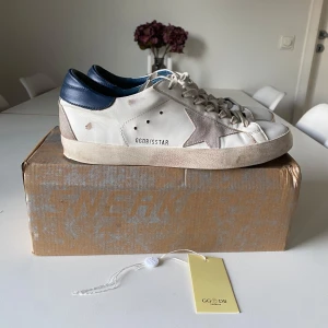 Golden Goose sneakers med stjärndetalj - Snygga Golden Goose sneakers i vitt med en grå stjärndetalj på sidan och mörkblå häl. Skorna har en klassisk snörning och en slitstark gummisula. Perfekta för en avslappnad stil. Frågor eller funderingar är det bara att höra av dig. Kanppt andävnda!!