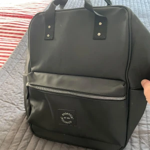 Svart ryggsäck från Essentials - Snygg och praktisk svart ryggsäck från Essentials med dragkedja framtill. Perfekt för vardagsbruk med justerbara axelremmar och ett stilrent utseende. Ryggsäcken har en fyrkantig form och är tillverkad i syntetmaterial.