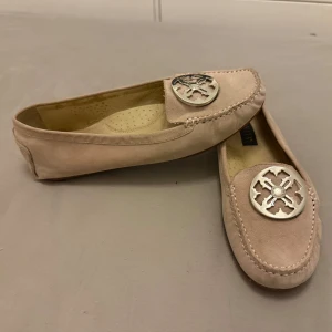 Beige/rosa mockaloafers från Novita - Snygga beige/rosa loafers i mocka från Novita med dekorativ metallapplikation på ovansidan. Skorna har en klassisk design med rundad tå och gummisula för extra komfort. Perfekta för en stilren look.