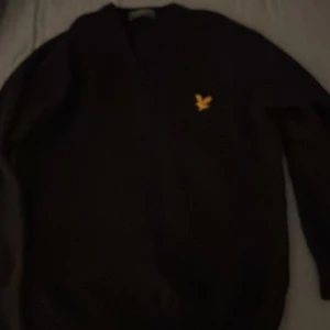 Svart tröja från Lyle & Scott - Säljer en stilren svart tröja från Lyle & Scott med deras ikoniska gula logga på bröstet. Tröjan har en v-ringad design och är perfekt för både vardag och mer uppklädda tillfällen.