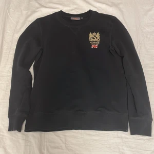 Morris Sweatshirt  - Snygg svart sweatshirt från Morris med broderad logga och brittisk flagga på bröstet. Tröjan har långa ärmar och en klassisk rund halsringning. Perfekt för en stilren och avslappnad look.
