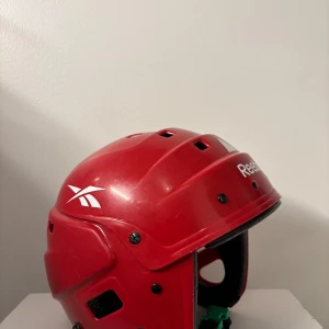 Hockey/skridskohjälm - Säljer en röd hjälm från Reebok med ventilationshål och justerbart spänne. Perfekt för sportaktiviteter där säkerhet är viktigt. Hjälmen har en stilren design med Reebok-loggan på sidan. Skridskohjälm i bra skick. Gör att ändra storlek med hjälp av skruvarna. Lite repig på utsidan mest av att ha legat i väskan. Fungerar bra som pulkahjälm/skridskohjälm/hockeyhjälm. Storlek 50-58 cm. 