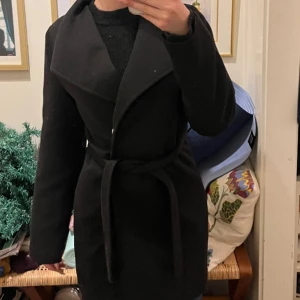 Svart kappa med knytband - Elegant svart kappa med knytband i midjan. Den har en klassisk design med långa ärmar och en stilren krage. Perfekt för kyligare dagar.