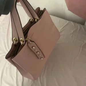Rosa handväska med pärldetaljer - Snygg helt ny rosa handväska från Frankrike med dekorativa pärlor på remmen. Väskan har två handtag och en dragkedja för säker förvaring. Perfekt för att bära dina dagliga nödvändigheter med stil. 