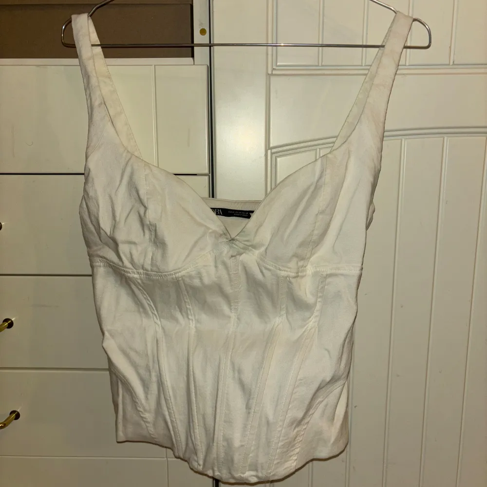 Säljer en stilren vit korsettopp från Zara. Toppen har breda axelband och en v-ringad design som ger en elegant look. Perfekt för att styla med jeans eller kjol för en chic outfit.. Yläosat.