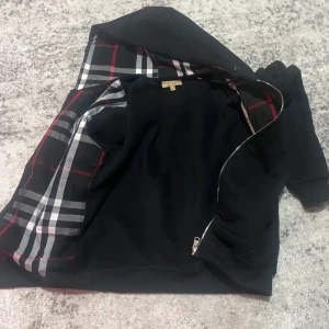 Svart burberry kofta - Snygg svart jacka med rutig insida i rött, vitt och grått. Jackan har en dragkedja och en broderad detalj på framsidan. Perfekt för en stilren look.